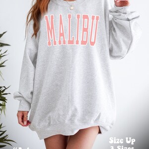 K&ouml;nnte beinhalten: Ein grauer Pullover mit dem Wort "MALIBU" in Pink auf der Vorderseite. Der Pullover ist oversized und hat eine lockere Passform. Das Model tr&auml;gt den Pullover eine Gr&ouml;&szlig;e gr&ouml;&szlig;er f&uuml;r einen oversized Look.