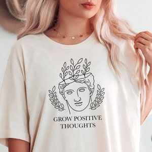 May include: White t-shirt with a black line drawing of a person's head with leaves growing out of it. The text "GROW POSITIVE THOUGHTS" is printed below the drawing.
