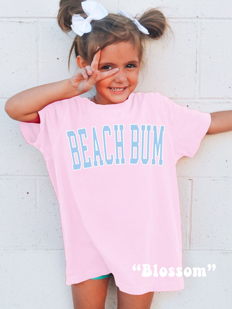 Pu&ograve; includere: Una bambina che indossa una maglietta rosa oversize con il testo "BEACH BUM" in lettere blu. La maglietta ha anche il testo "Blossom" in lettere bianche in basso.
