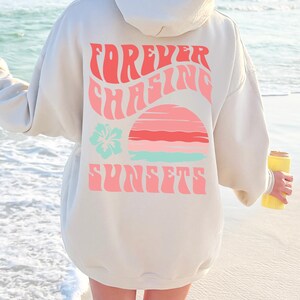 Pode incluir: Uma camisola com capuz bege com um gr&aacute;fico nas costas com o texto "FOREVER CHASING SUNSETS" em letras cor-de-rosa onduladas. O design inclui um gr&aacute;fico estilizado de um p&ocirc;r do sol em rosa e azul-claro e um &iacute;cone de flor de hibisco azul-claro.