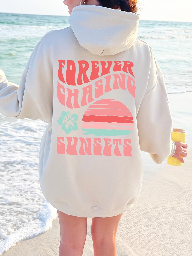 Pode incluir: Uma camisola com capuz bege com um gr&aacute;fico nas costas com o texto "FOREVER CHASING SUNSETS" em letras cor-de-rosa onduladas. O design inclui um gr&aacute;fico estilizado de um p&ocirc;r do sol em rosa e azul-claro e um &iacute;cone de flor de hibisco azul-claro.