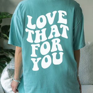 K&ouml;nnte beinhalten: Ein t&uuml;rkisfarbenes T-Shirt mit dem Text "LOVE THAT FOR YOU" in wei&szlig;er, retro-artiger Schrift. Das Shirt wird an einem Model gezeigt und die Farbe wird als "Seafoam" beschrieben.