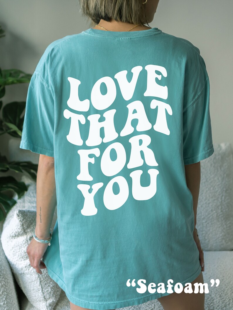 K&ouml;nnte beinhalten: Ein t&uuml;rkisfarbenes T-Shirt mit dem Text "LOVE THAT FOR YOU" in wei&szlig;er, retro-artiger Schrift. Das Shirt wird an einem Model gezeigt und die Farbe wird als "Seafoam" beschrieben.
