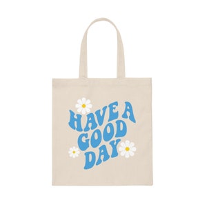 May include: A beige canvas tote bag with blue text that reads "HAVE A GOOD DAY" in a retro font. The text is surrounded by white daisies.