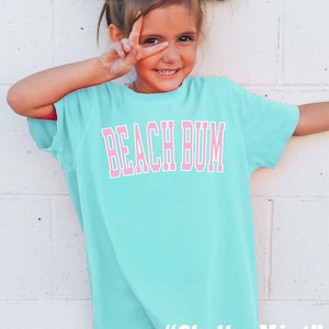 May include: A light turquoise t-shirt with the words "BEACH BUM" in pink block letters. The shirt is worn by a young girl with white bows in her hair. The text "Chalky Mint" is printed at the bottom of the shirt.