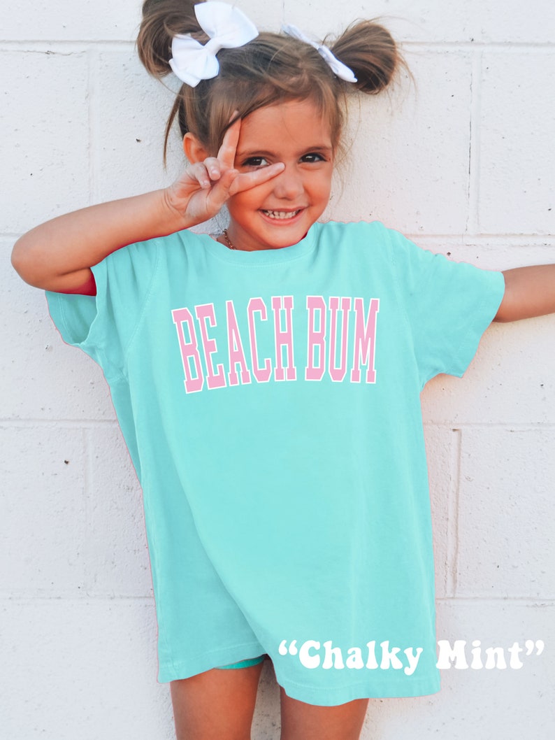 May include: A light turquoise t-shirt with the words "BEACH BUM" in pink block letters. The shirt is worn by a young girl with white bows in her hair. The text "Chalky Mint" is printed at the bottom of the shirt.