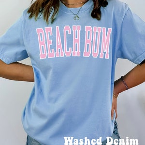 May include: A light blue t-shirt with the text "Beach Bum" in pink lettering. The shirt is being worn by a person wearing denim shorts. The text "Washed Denim" is visible at the bottom of the image.