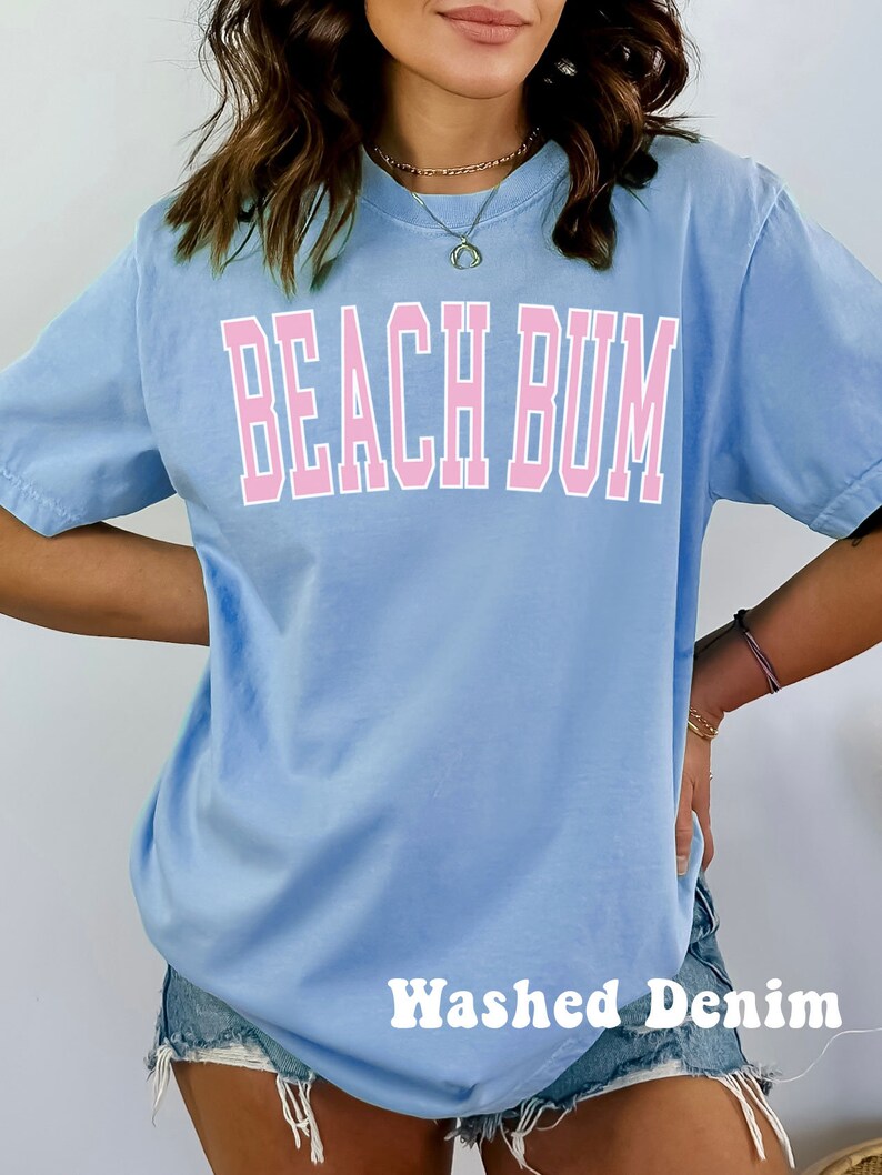 May include: A light blue t-shirt with the text "Beach Bum" in pink lettering. The shirt is being worn by a person wearing denim shorts. The text "Washed Denim" is visible at the bottom of the image.