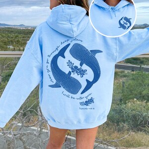 K&ouml;nnte beinhalten: Ein hellblauer Kapuzenpullover mit einer dunkelblauen Grafik auf der R&uuml;ckseite, die zwei Walhaie, florale Akzente und den Text "when you go through deep waters I will be with you. Isaiah 43:2" zeigt. Ein kleines passendes Logo ist vorne.