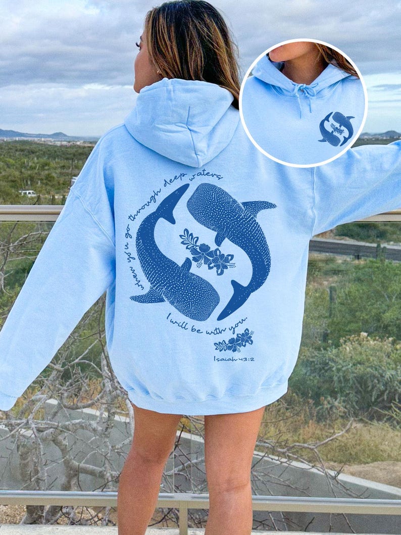K&ouml;nnte beinhalten: Ein hellblauer Kapuzenpullover mit einer dunkelblauen Grafik auf der R&uuml;ckseite, die zwei Walhaie, florale Akzente und den Text "when you go through deep waters I will be with you. Isaiah 43:2" zeigt. Ein kleines passendes Logo ist vorne.