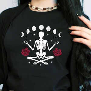 May include: Black t-shirt with a white skeleton sitting in a yoga pose with a crescent moon and stars around it. There are two red roses on either side of the skeleton.