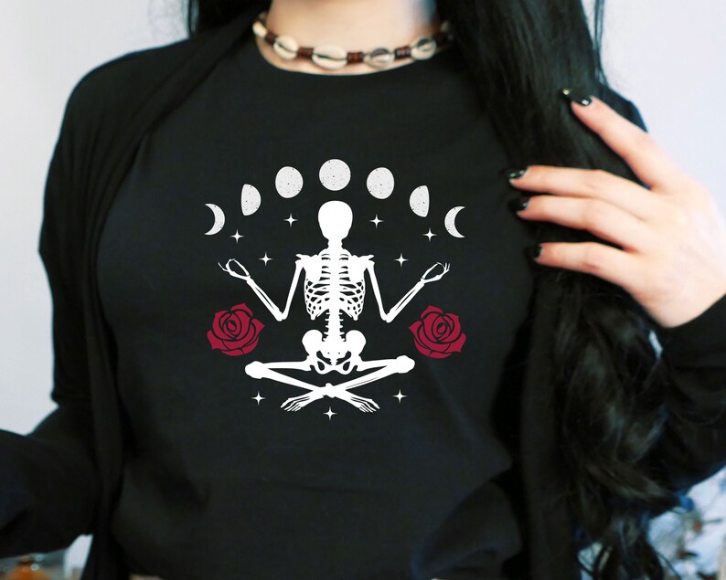 May include: Black t-shirt with a white skeleton sitting in a yoga pose with a crescent moon and stars around it. There are two red roses on either side of the skeleton.