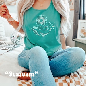 K&ouml;nnte beinhalten: Ein t&uuml;rkisfarbenes Tanktop mit einem wei&szlig;en Wal- und Sonnenmotiv. Der Text "Seafoam" steht unter dem Motiv.