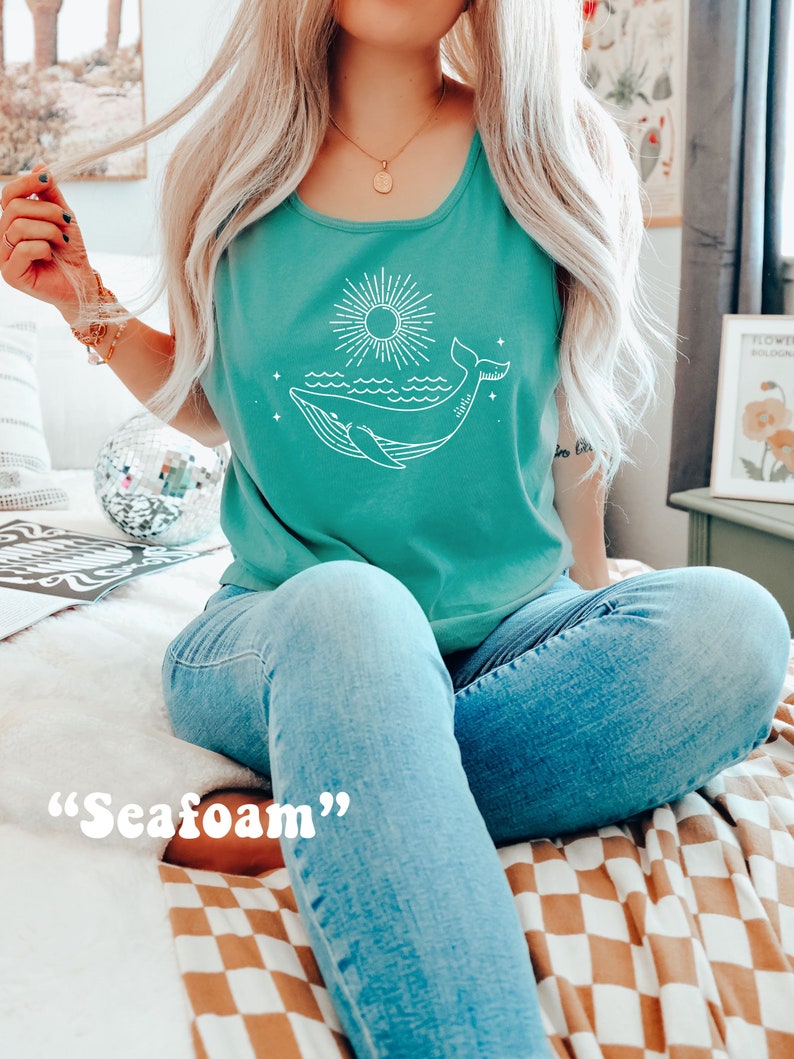 K&ouml;nnte beinhalten: Ein t&uuml;rkisfarbenes Tanktop mit einem wei&szlig;en Wal- und Sonnenmotiv. Der Text "Seafoam" steht unter dem Motiv.