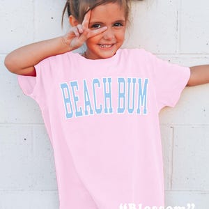 May include: A light pink t-shirt with the words "BEACH BUM" in light blue block letters. The word "Blossom" is printed in white script at the bottom of the shirt. The shirt is worn by a young girl with white bows in her hair.