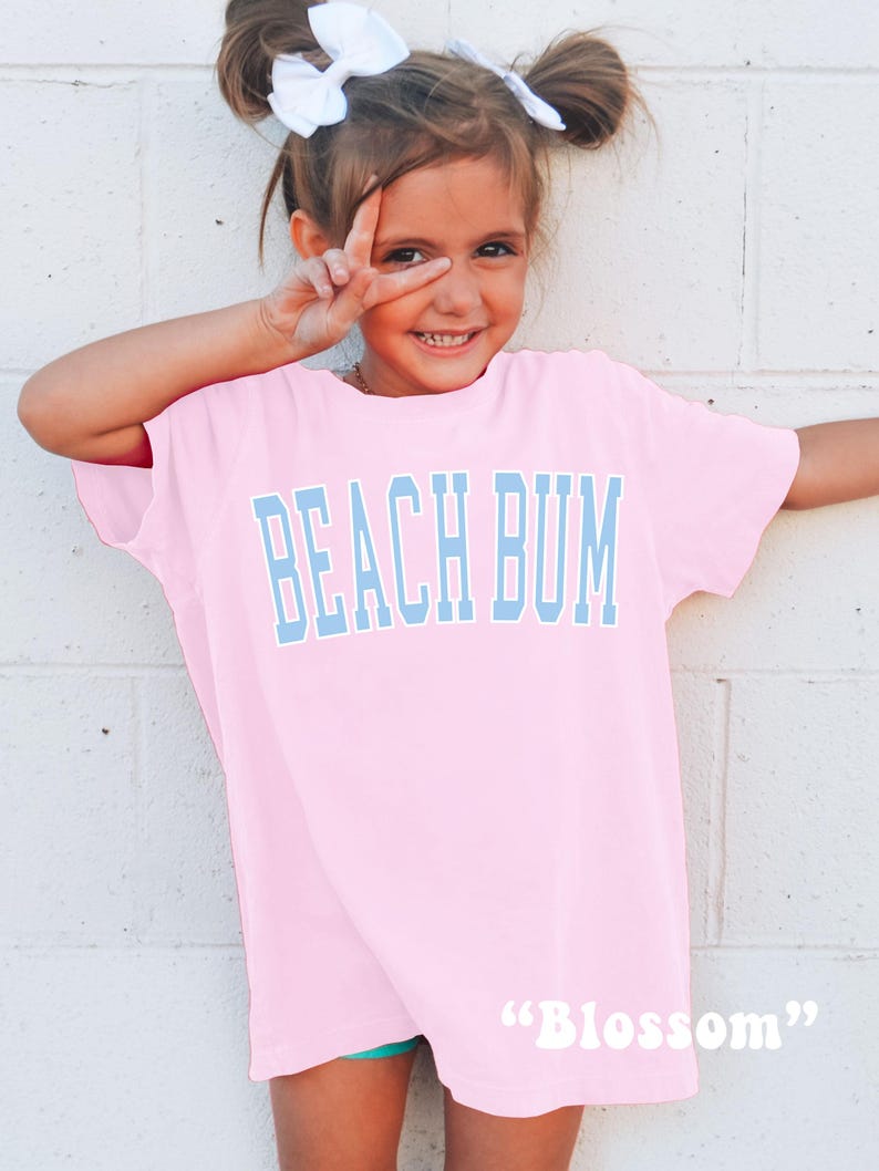 May include: A light pink t-shirt with the words "BEACH BUM" in light blue block letters. The word "Blossom" is printed in white script at the bottom of the shirt. The shirt is worn by a young girl with white bows in her hair.