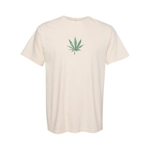 以下が含まれることがあります： 緑色のマリファナの葉のグラフィックが描かれたクリーム色のTシャツ