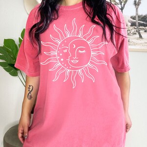 K&ouml;nnte beinhalten: Ein rosafarbenes Oversize-T-Shirt mit einem wei&szlig;en Sonnen- und Mondmotiv. Die Sonne hat ein Gesicht und der Mond ist halbmondf&ouml;rmig.