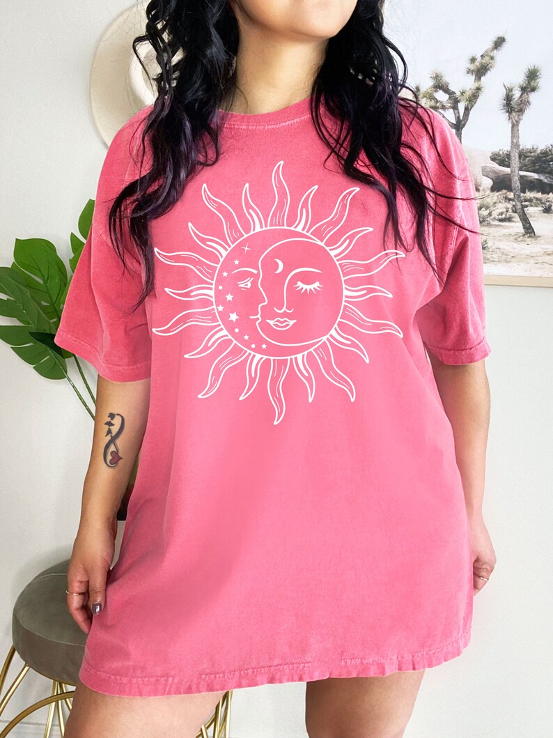 K&ouml;nnte beinhalten: Ein rosafarbenes Oversize-T-Shirt mit einem wei&szlig;en Sonnen- und Mondmotiv. Die Sonne hat ein Gesicht und der Mond ist halbmondf&ouml;rmig.