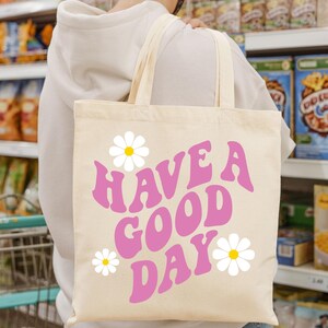 May include: A beige tote bag with pink retro-style lettering that says "Have a Good Day" and white daisy flowers.