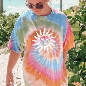May include: A tie-dye t-shirt in shades of blue, green, pink, and orange. The shirt features a white sun graphic with a face in the center. The shirt is a casual, oversized fit.