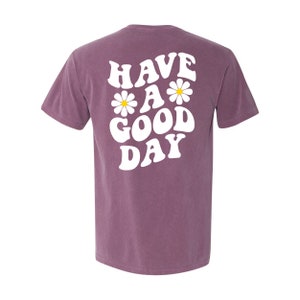May include: A purple t-shirt with the text "HAVE A GOOD DAY" in white with daisy flowers on either side of the "A".