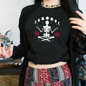 May include: A black cropped t-shirt with a white graphic of a skeleton meditating with a crescent moon and stars. The skeleton is surrounded by six moons in different phases. Two red roses are on either side of the skeleton.
