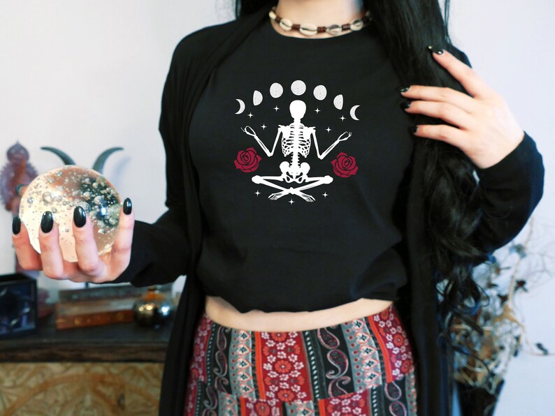 May include: A black cropped t-shirt with a white graphic of a skeleton meditating with a crescent moon and stars. The skeleton is surrounded by six moons in different phases. Two red roses are on either side of the skeleton.