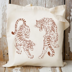 May include: A white tote bag with two brown tiger illustrations. The tigers are facing opposite directions and are drawn in a simple, graphic style.