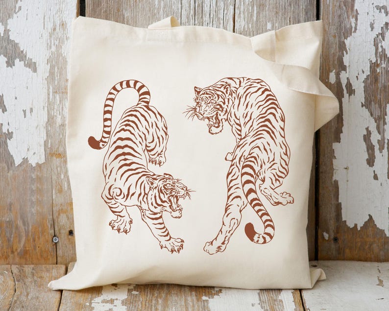 May include: A white tote bag with two brown tiger illustrations. The tigers are facing opposite directions and are drawn in a simple, graphic style.