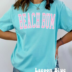 May include: A light blue t-shirt with the text "Beach Bum" in pink lettering. The shirt is shown on a model wearing denim shorts. The text "Lagoon Blue" is written in white at the bottom of the image.