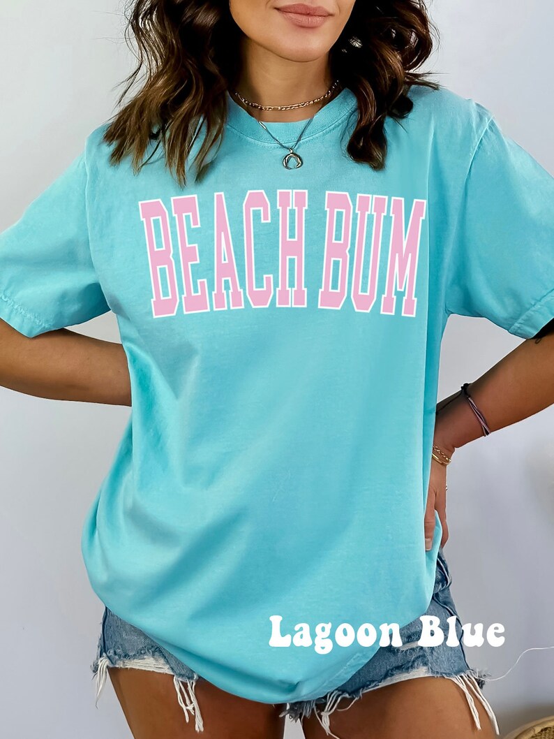 May include: A light blue t-shirt with the text "Beach Bum" in pink lettering. The shirt is shown on a model wearing denim shorts. The text "Lagoon Blue" is written in white at the bottom of the image.