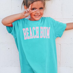 May include: A turquoise t-shirt with the words "BEACH BUM" in pink block letters. The shirt is worn by a young person with white bows in their hair, making a peace sign with their fingers.