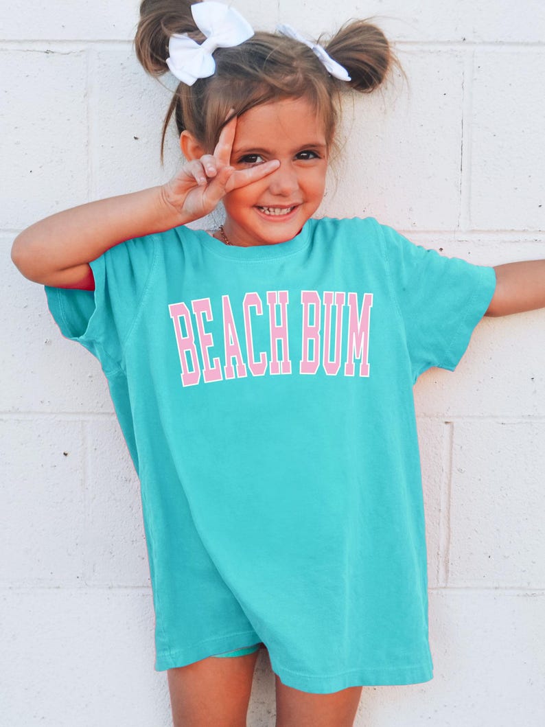 May include: A turquoise t-shirt with the words "BEACH BUM" in pink block letters. The shirt is worn by a young person with white bows in their hair, making a peace sign with their fingers.