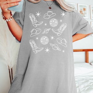 Puede incluir: Una camiseta gris de gran tama&ntilde;o con un dise&ntilde;o gr&aacute;fico blanco que presenta botas de vaquero, sombreros, bolas de discoteca y estrellas.