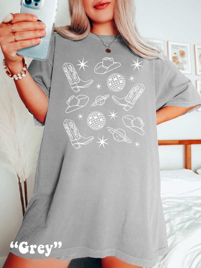 Puede incluir: Una camiseta gris de gran tama&ntilde;o con un dise&ntilde;o gr&aacute;fico blanco que presenta botas de vaquero, sombreros, bolas de discoteca y estrellas.