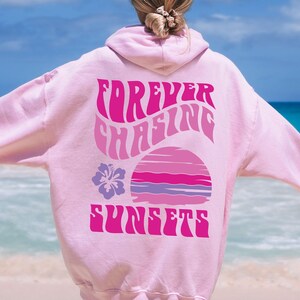 Pode incluir: Uma camisola com capuz cor-de-rosa claro com um gr&aacute;fico nas costas com o texto "FOREVER CHASING SUNSETS" numa fonte ondulada. O design inclui um p&ocirc;r do sol estilizado em tons de rosa e roxo, acompanhado por uma flor de hibisco roxa.