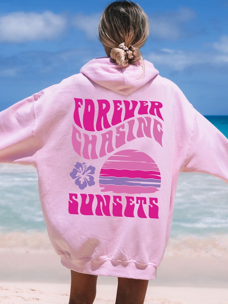 Pode incluir: Uma camisola com capuz cor-de-rosa claro com um gr&aacute;fico nas costas com o texto "FOREVER CHASING SUNSETS" numa fonte ondulada. O design inclui um p&ocirc;r do sol estilizado em tons de rosa e roxo, acompanhado por uma flor de hibisco roxa.