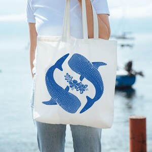 May include: A white tote bag with a blue design of two whale sharks swimming in a circle with a floral design in the center.