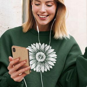 Pu&ograve; includere: Una donna indossa una felpa verde scuro con un motivo di girasole bianco. Tiene uno smartphone in mano e ascolta musica con cuffie bianche.
