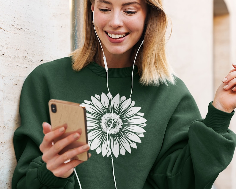 Pu&ograve; includere: Una donna indossa una felpa verde scuro con un motivo di girasole bianco. Tiene uno smartphone in mano e ascolta musica con cuffie bianche.