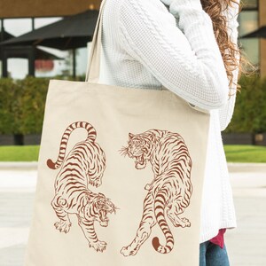 May include: A beige tote bag with two brown tigers printed on it. The tigers are facing each other and have a fierce expression. The bag is being held by a person wearing a white sweater.