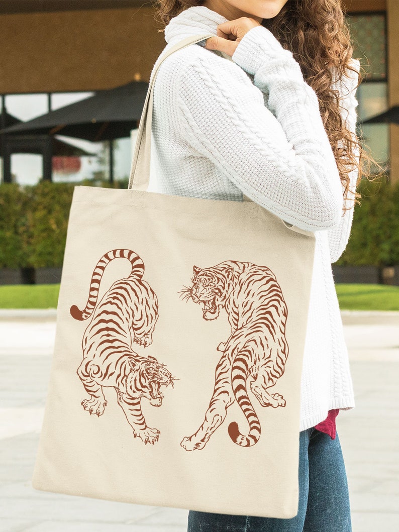 May include: A beige tote bag with two brown tigers printed on it. The tigers are facing each other and have a fierce expression. The bag is being held by a person wearing a white sweater.