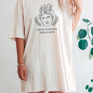 May include: A white oversized t-shirt with a black line drawing of a face with leaves growing out of the top of the head. The text "GROW POSITIVE THOUGHTS" is printed below the drawing.