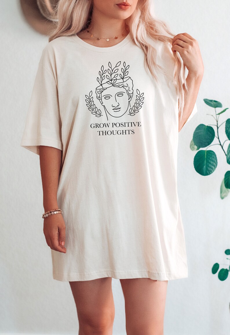 May include: A white oversized t-shirt with a black line drawing of a face with leaves growing out of the top of the head. The text "GROW POSITIVE THOUGHTS" is printed below the drawing.