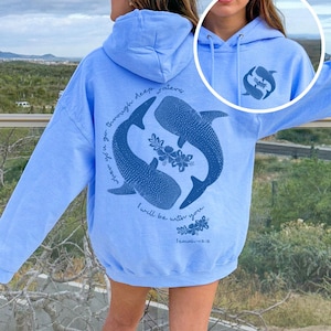 K&ouml;nnte beinhalten: Ein hellblauer Kapuzenpullover mit einer dunkelblauen Grafik auf der R&uuml;ckseite, die zwei Walhaie in einem Kreis, florale Akzente und den Text "when you go through deep waters I will be with you. Isaiah 43:2" zeigt. Vorne befindet sich ein kleines passendes Logo.