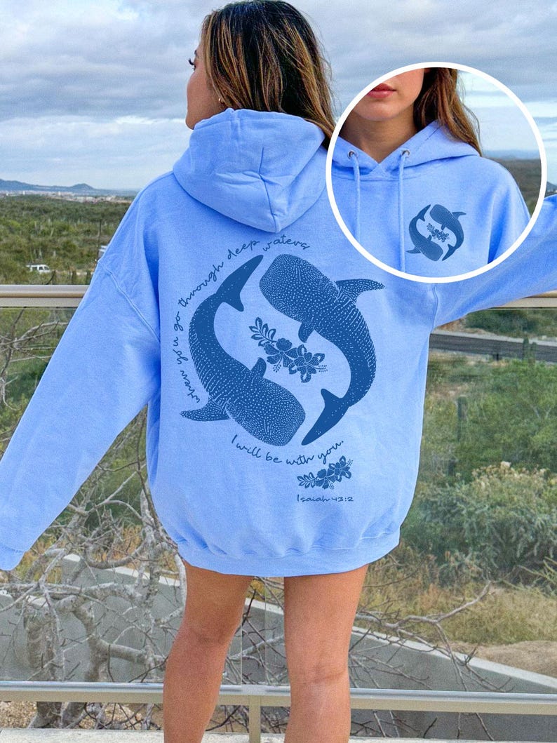 K&ouml;nnte beinhalten: Ein hellblauer Kapuzenpullover mit einer dunkelblauen Grafik auf der R&uuml;ckseite, die zwei Walhaie in einem Kreis, florale Akzente und den Text "when you go through deep waters I will be with you. Isaiah 43:2" zeigt. Vorne befindet sich ein kleines passendes Logo.
