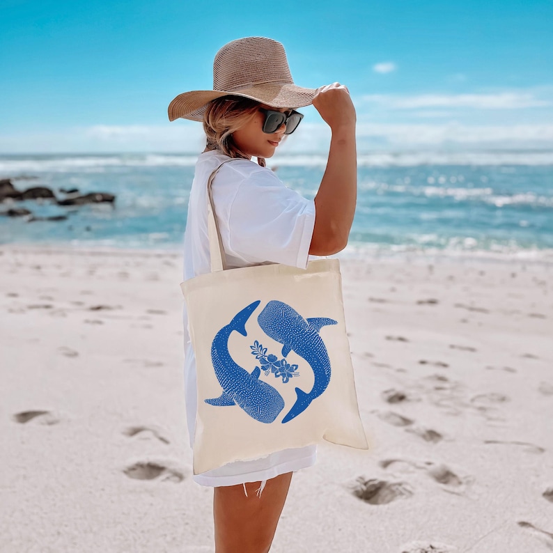May include: A woman wearing a straw hat and sunglasses is carrying a white tote bag with a blue whale shark design. The bag has a floral design in the center.