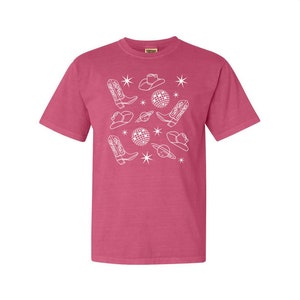 Puede incluir: Una camiseta rosa con un dise&ntilde;o blanco de botas de vaquero, sombreros, bolas de discoteca y estrellas.