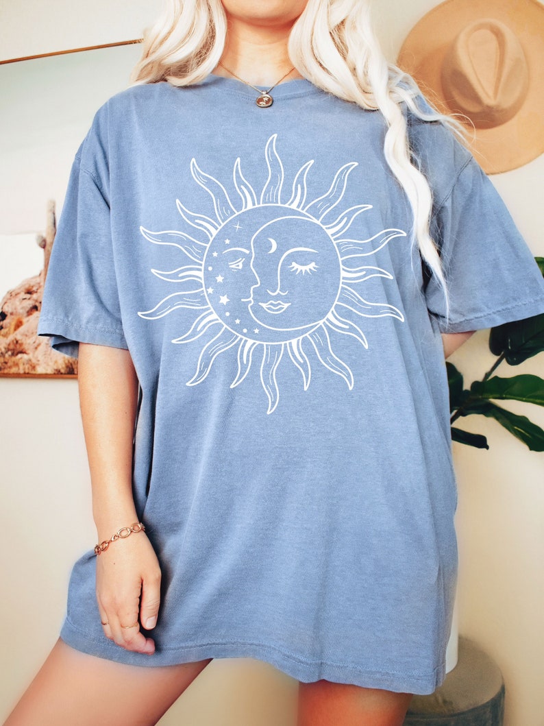 K&ouml;nnte beinhalten: Ein hellblaues Oversize-T-Shirt mit einem wei&szlig;en Sonnen- und Mondmotiv. Sonne und Mond haben Gesichter und sind von Sternen umgeben.